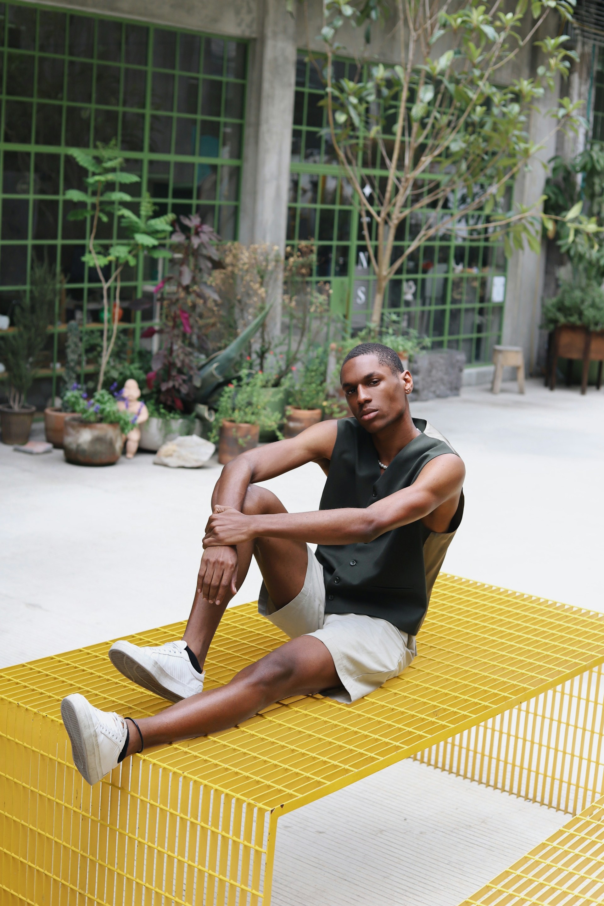 A man sits on a bright yellow metal grate bench in an open-air industrial space. He is wearing a sleeveless dark green top and light-colored shorts, with potted plants and large windows in the background.