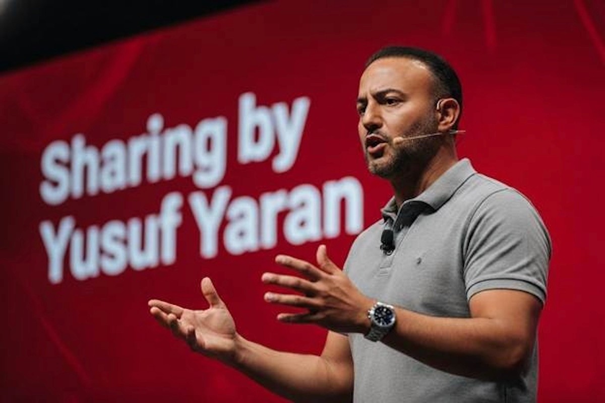 Yusuf Yaran speaking on stage before red presentation screen during hospitality industry event.