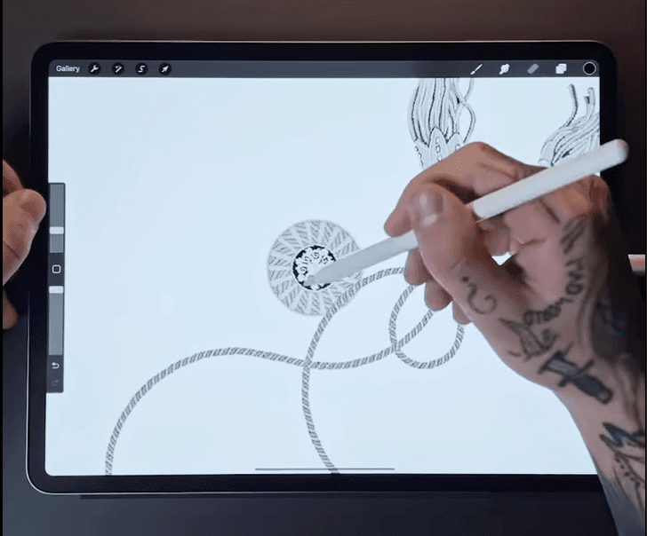 A person using a digital pen to draw intricate designs on a tablet, focusing on circular patterns and rope-like elements. The person's hand has visible tattoos, including a cross and other small symbols.