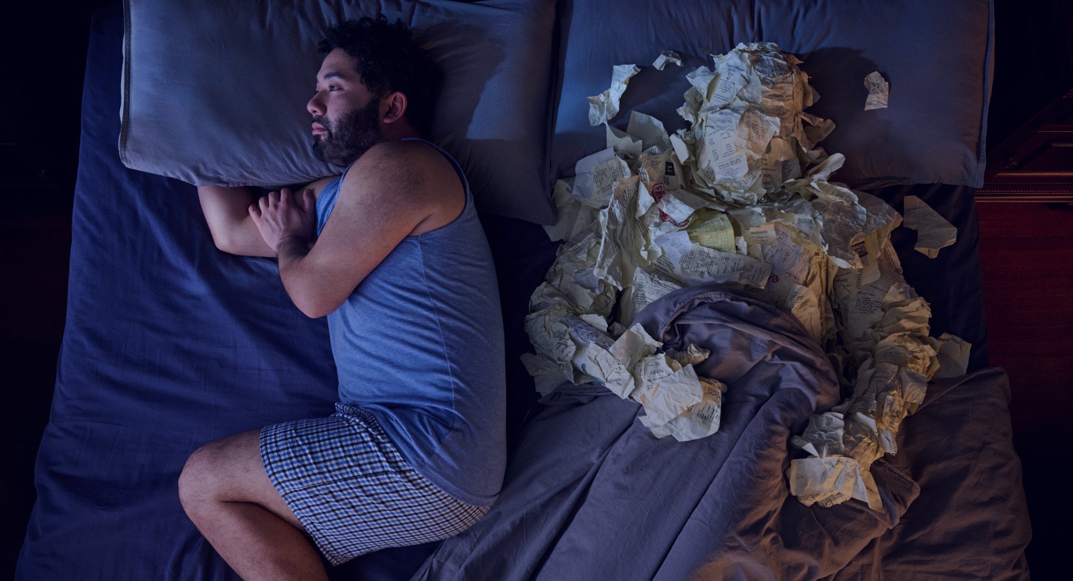 A man sleeps in bed next to a humanoid figure, both under sheets in a dimly lit room.