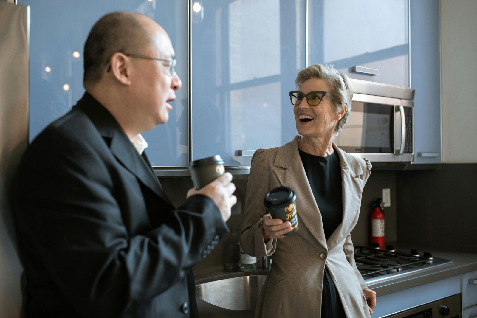 Two people standing in a modern kitchen, conversing while holding coffee cups; one wears a dark suit and glasses, the other a light blazer and glasses, with blue cabinets, a microwave, stove, and a fire extinguisher visible in the background.