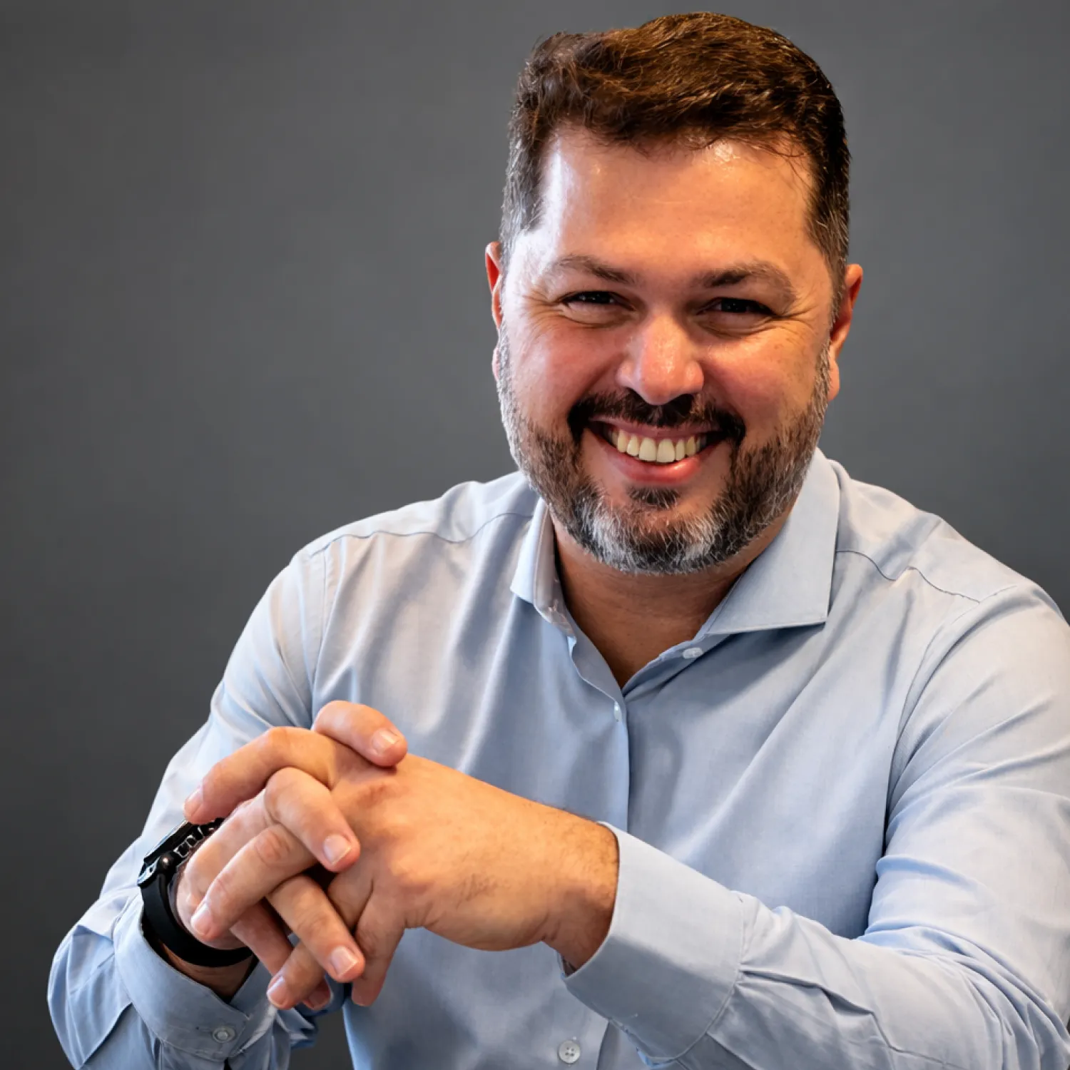 A young person with curly hair smiles at the camera while sitting in front of a laptop against a dark background.