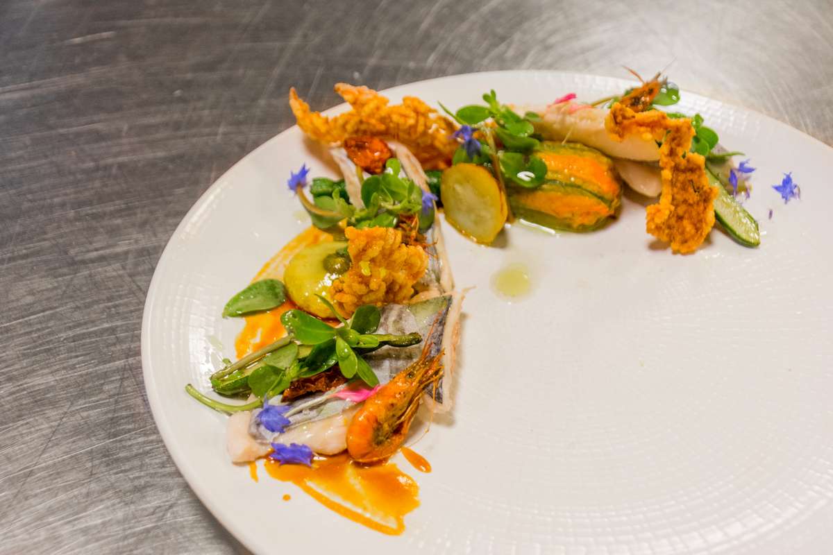 Three cooked langoustines served on a white plate with lime wedges, microgreens, and small purple flowers.