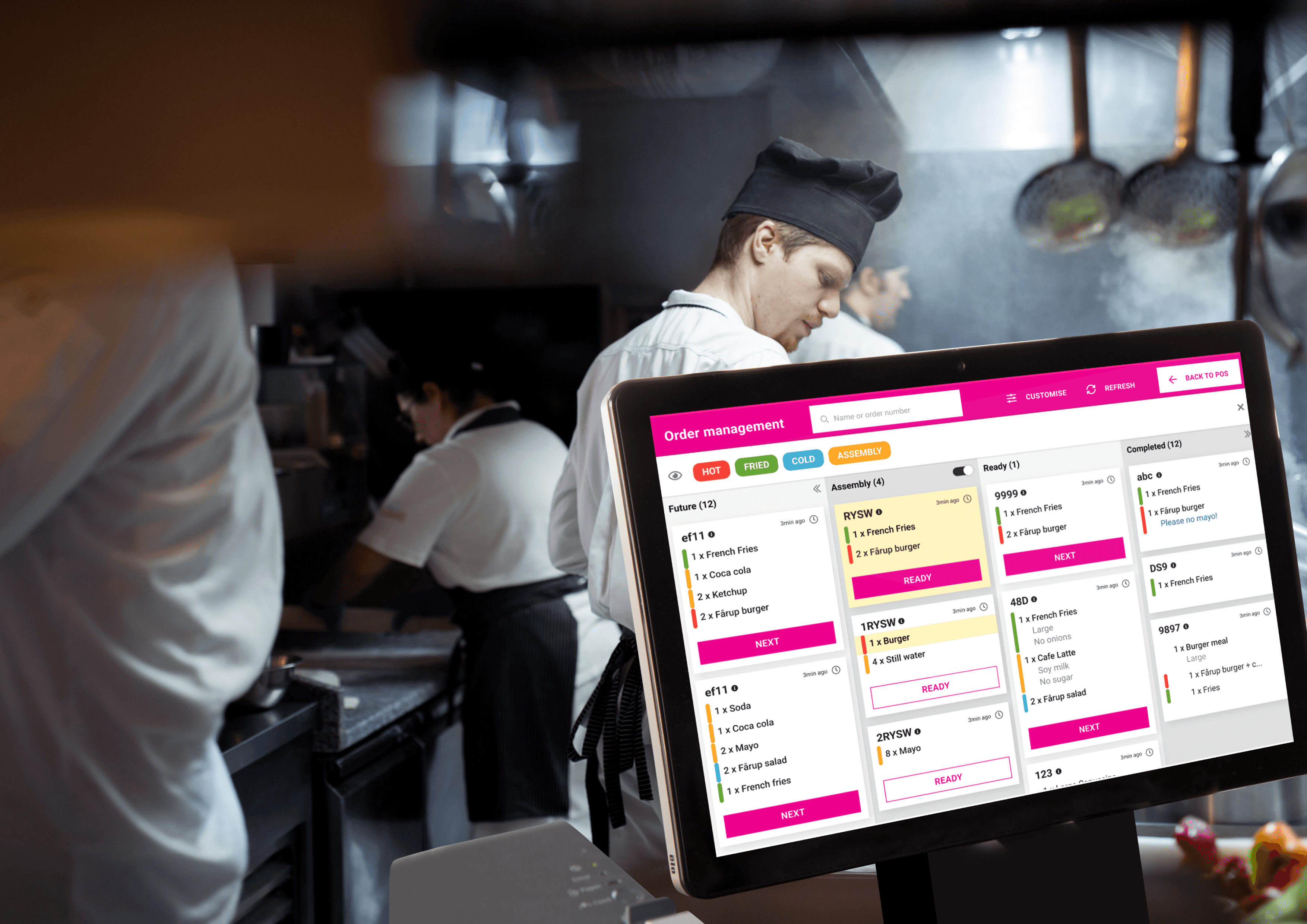 A young person with curly hair smiles at the camera while sitting in front of a laptop against a dark background.