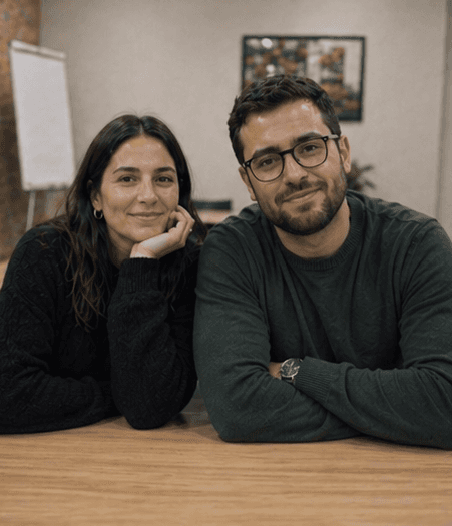 Dos mujeres jóvenes estudiando juntas en una mesa iluminada por luz natural. Una de ellas escribe mientras la otra observa con atención, creando un ambiente de colaboración y aprendizaje.
