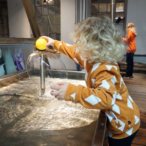 A child with curly hair plays with a water table, using a yellow ball. Another child in an orange shirt is visible in the background.