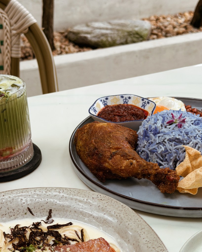 A close-up of a plate featuring fried chicken drumstick, blue rice garnished with a flower, sambal in a small bowl, and crispy snacks, accompanied by a green drink in a glass.