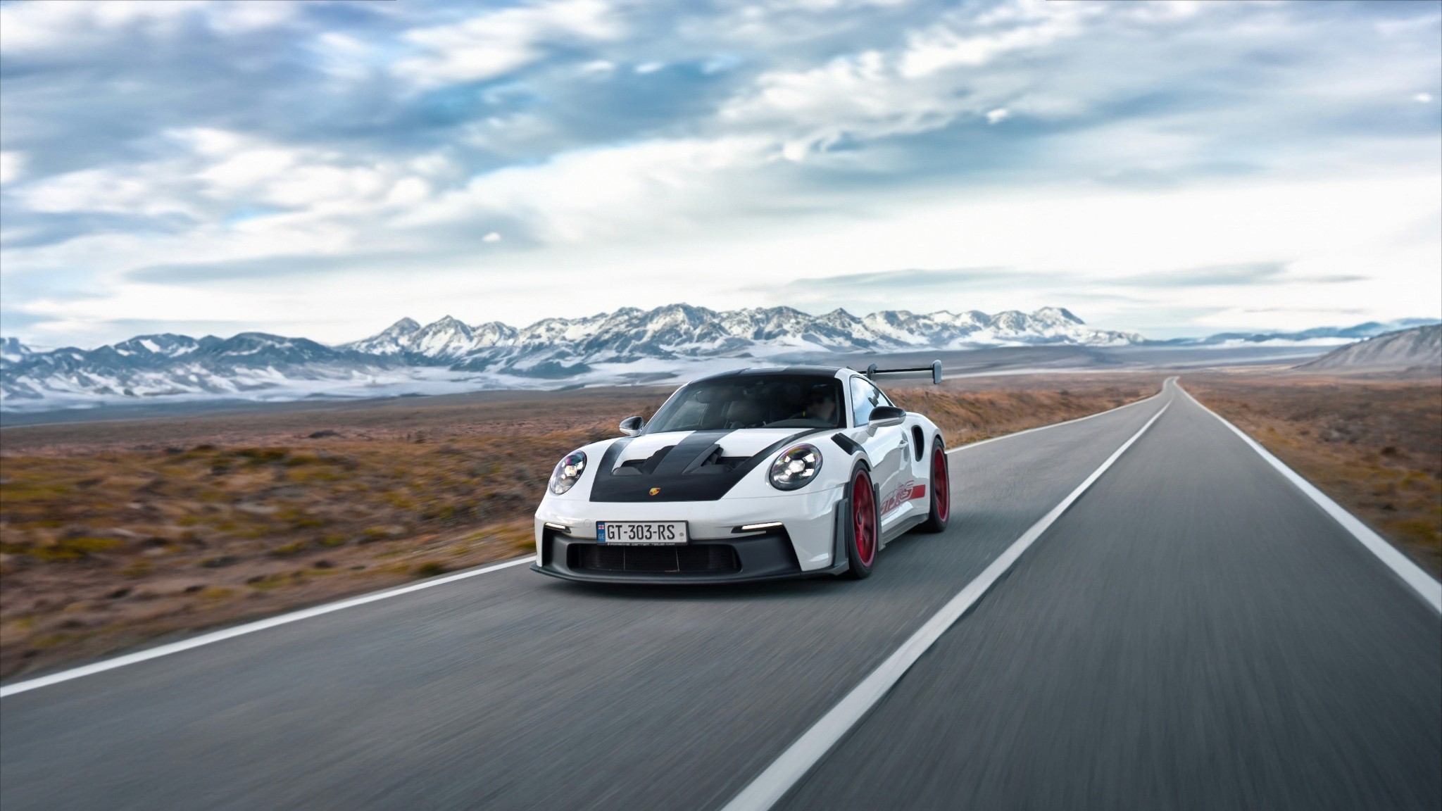 A sleek sports car with a white body and black accents speeds down an open highway, set against a backdrop of vast, rocky desert and distant snow-capped mountains under a cloudy sky.
