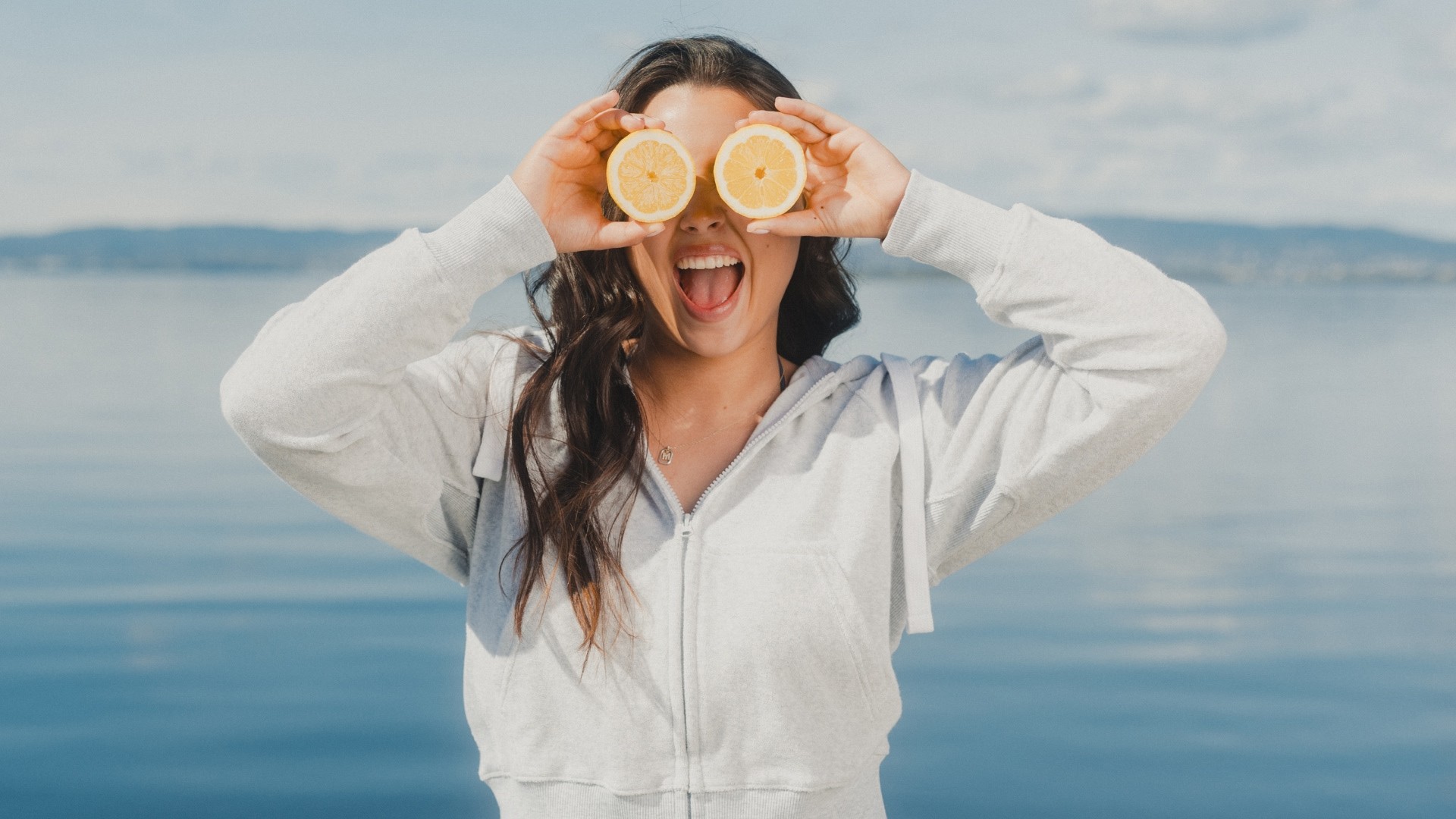 Young woman dressed in light grey ziphood, holding two lemons in front of her eyes and smiling