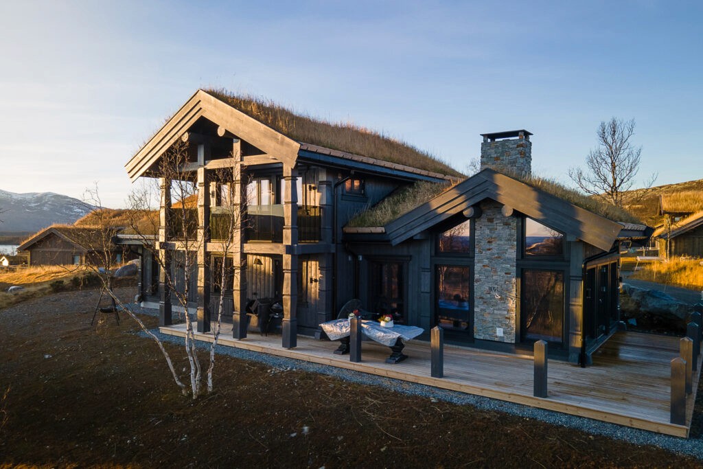 A dark post-and-beam house with a stone chimney and large living room windows, blending rustic charm with modern elegance against a natural backdrop