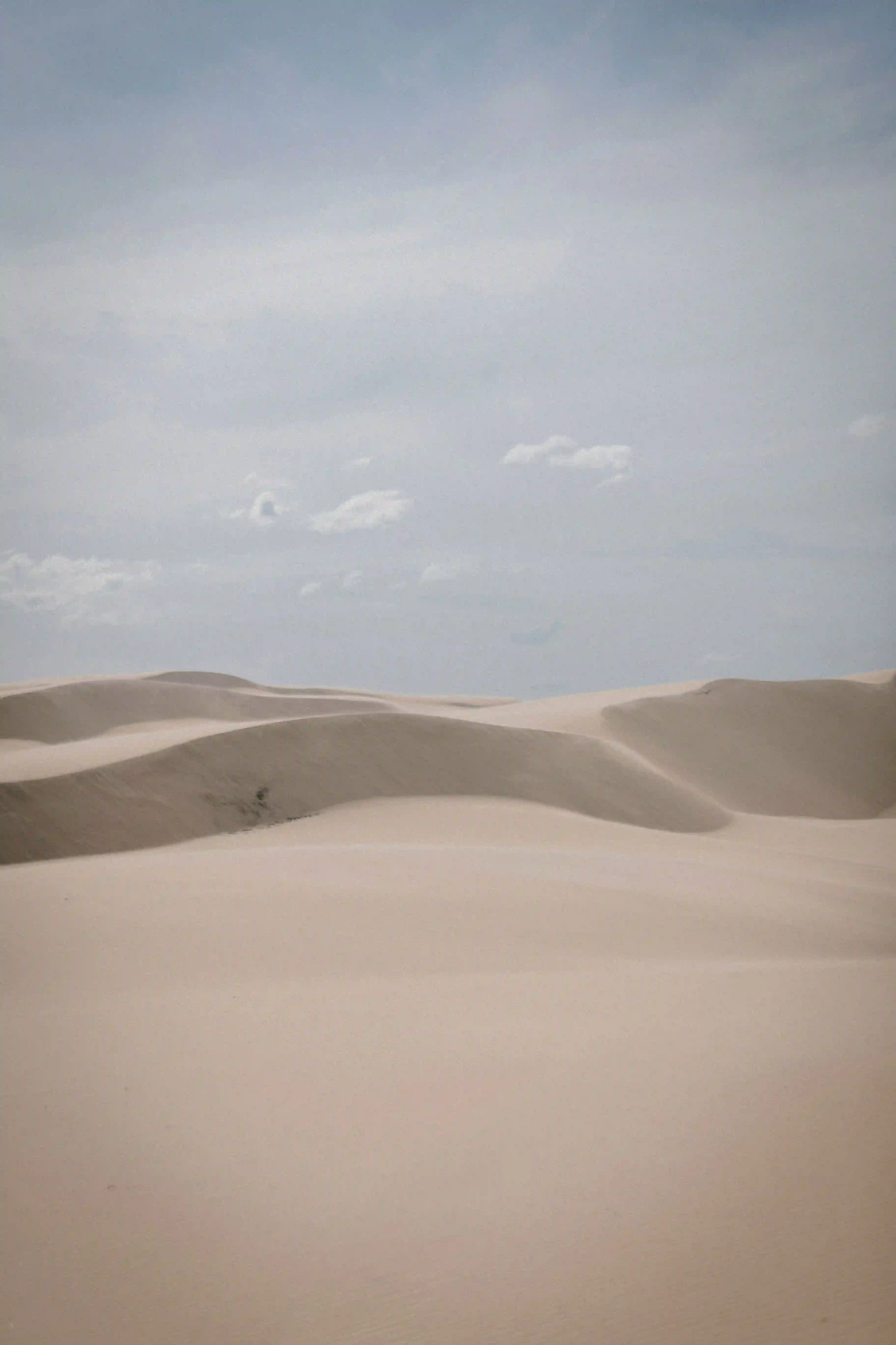 A lone person walking across large sand dunes in a vast desert landscape under a cloudy sky