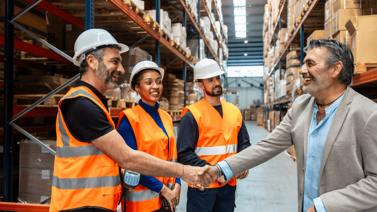 Warehouse manager shaking hands with a client in front of stocked pallet racking.