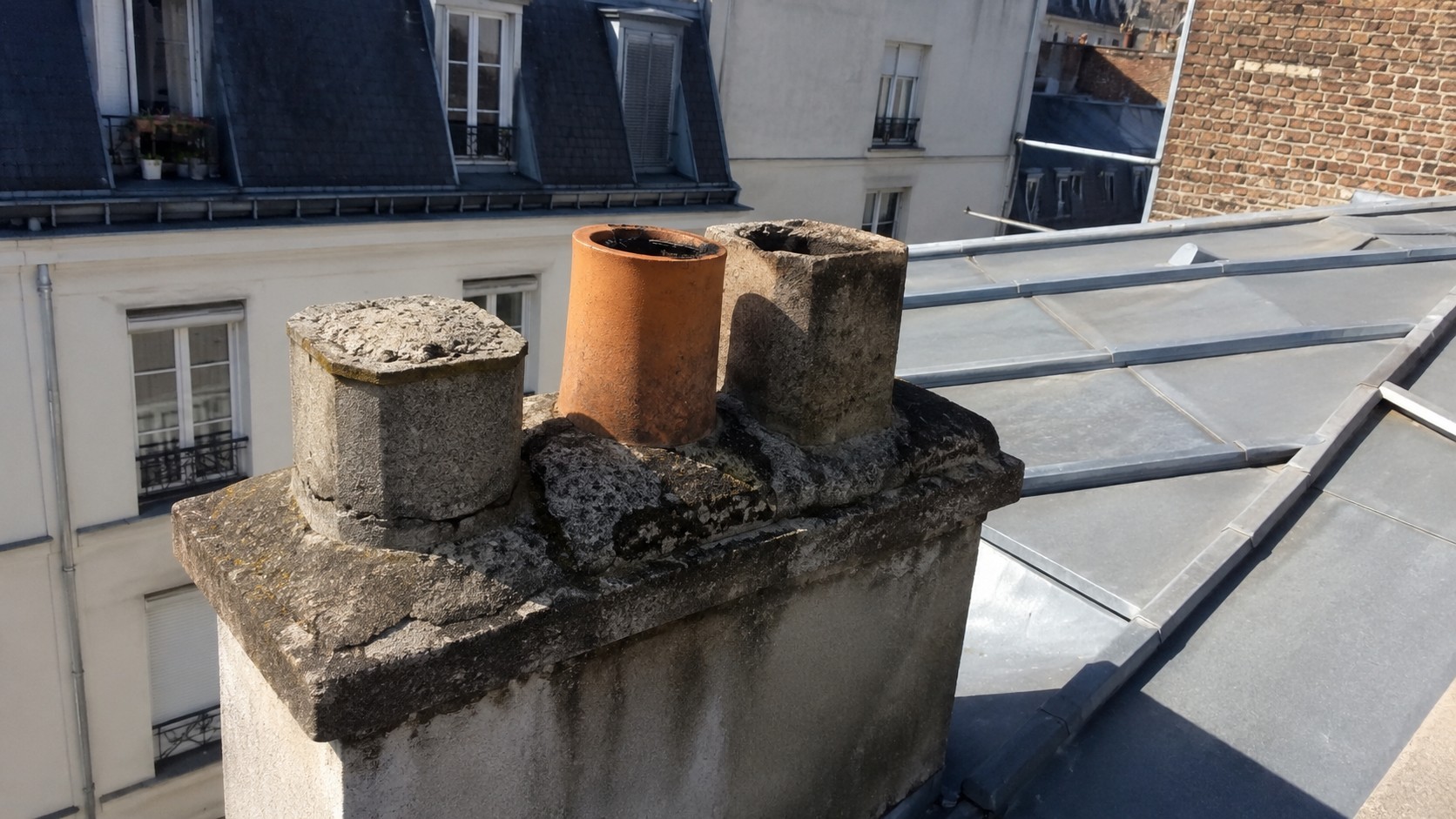 Ancien conduit de cheminée non utilisé sur une toiture parisienne, avec souche maçonnée vieillie, conduit partiellement obturé et couverture en zinc exposée au soleil.