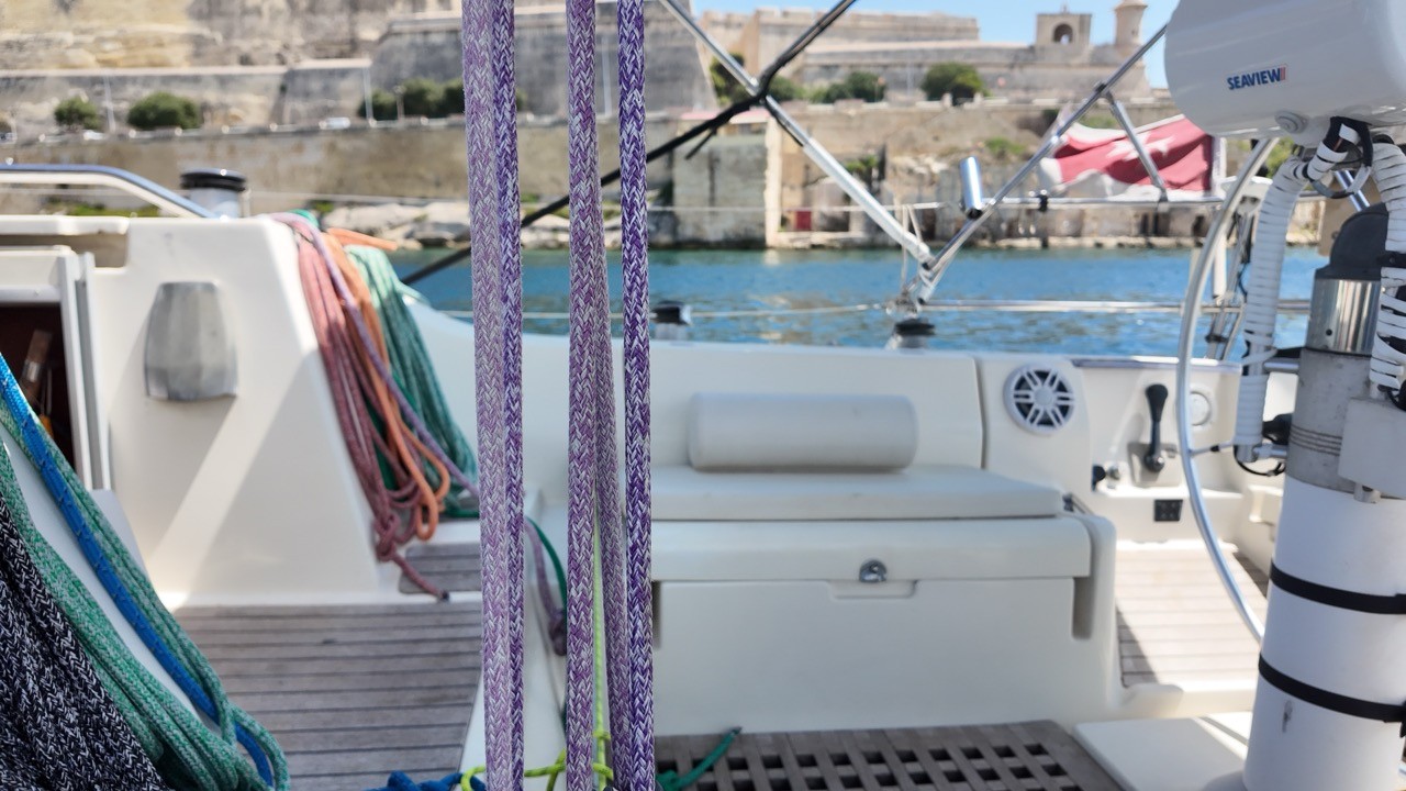 Close-up of the Beneteau First 38.5's cockpit showing coiled mooring lines, a cushioned seat, and a Seaview satellite dome, with a historic stone fortress and harbor visible beyond the rail.