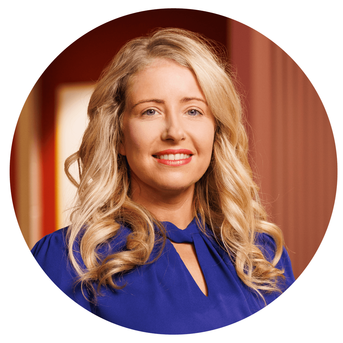 A circular, studio‑style head‑and‑shoulders portrait of a person with long, wavy blonde hair, wearing a royal blue blouse with a gathered neckline. The background features warm indoor tones, creating a professional and polished profile image.