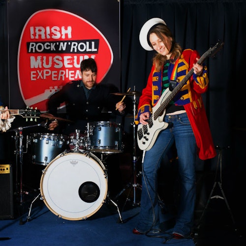 A person in colorful, band-style attire plays a white bass guitar next to a drummer, with an "Irish Rock 'N' Roll Museum Experience" banner behind.