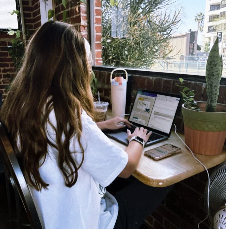 A person wearing headphones sits at a desk with a large curved monitor displaying graphic design software, surrounded by a laptop, smartphone, and potted plant, creating a modern workspace setup.