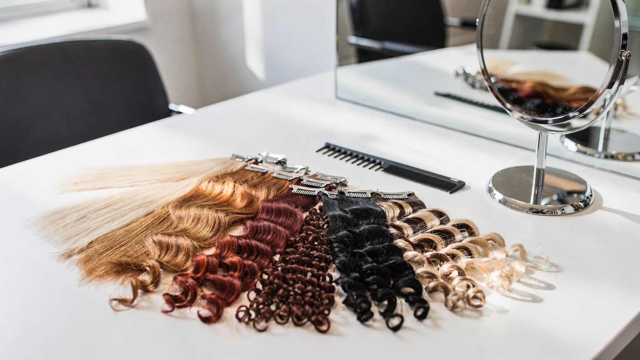 A salon consultation table with hair extension swatches in different curl patterns and colors arranged beside a comb, clips, and mirror.