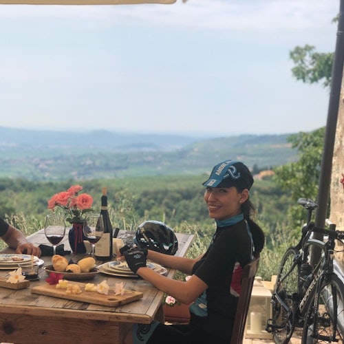 Una mujer con ropa de ciclismo se sienta en una mesa al aire libre con comida, vino y flores, con un paisaje colinoso y pintoresco de fondo.