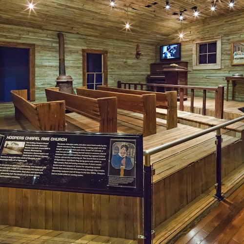 Interiér dřevěné kaple s lavicemi, kamny, historickou informační tabulí, televizí a pianem. Cedule s nápisem "Hoopers Chapel AME Church."