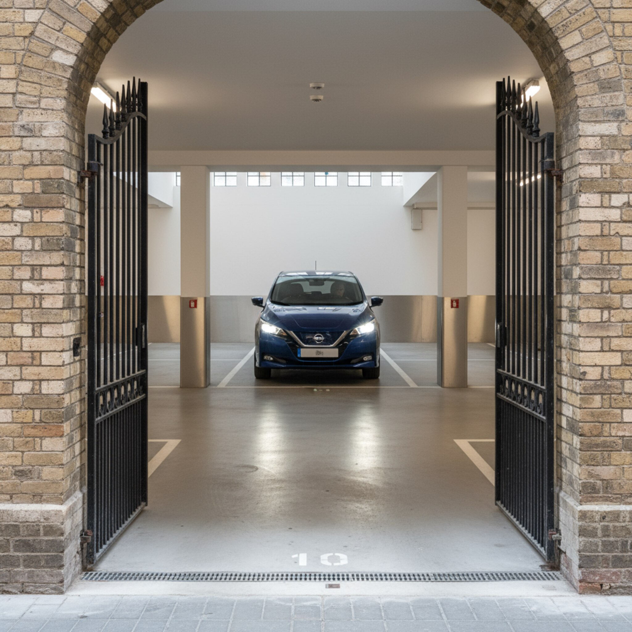 Ein schmales Altbau-Tor öffnet sich zu einer sauberen Innenhofgarage. Scheinwerfer fangen die weiße Linienführung und sanft glänzende Betonflächen ein. Ein Kleinwagen rollt langsam hinein, die Umgebung ist aufgeräumt, trocken, sicher. Die klare Geometrie der Stellplatznummern dominiert die reduzierte, hochwertige Szene.