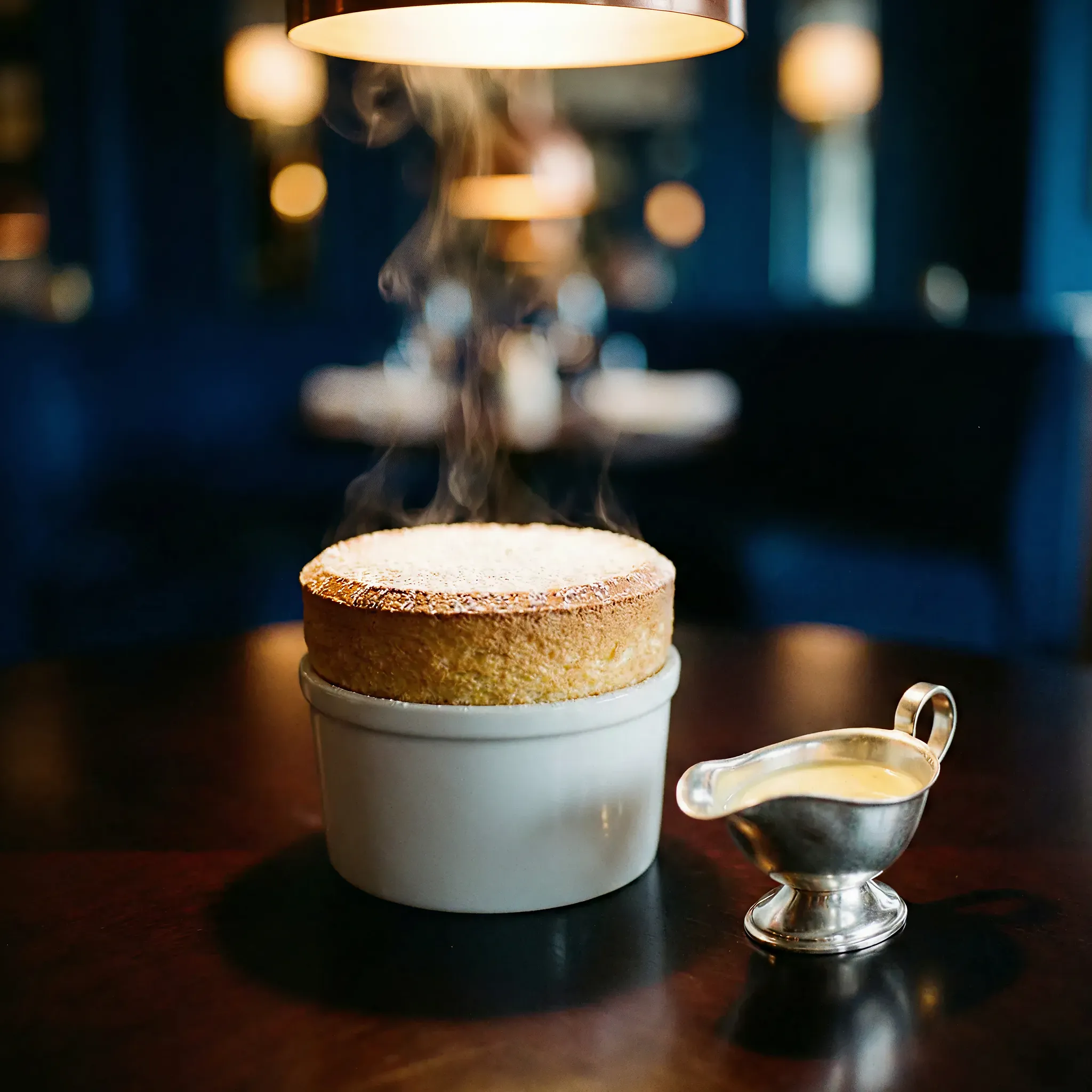 A warm soufflé dusted with powdered sugar emits steam in a white ramekin under a glowing lamp. A silver sauceboat beside it on a dark table creates a cozy, elegant ambiance.