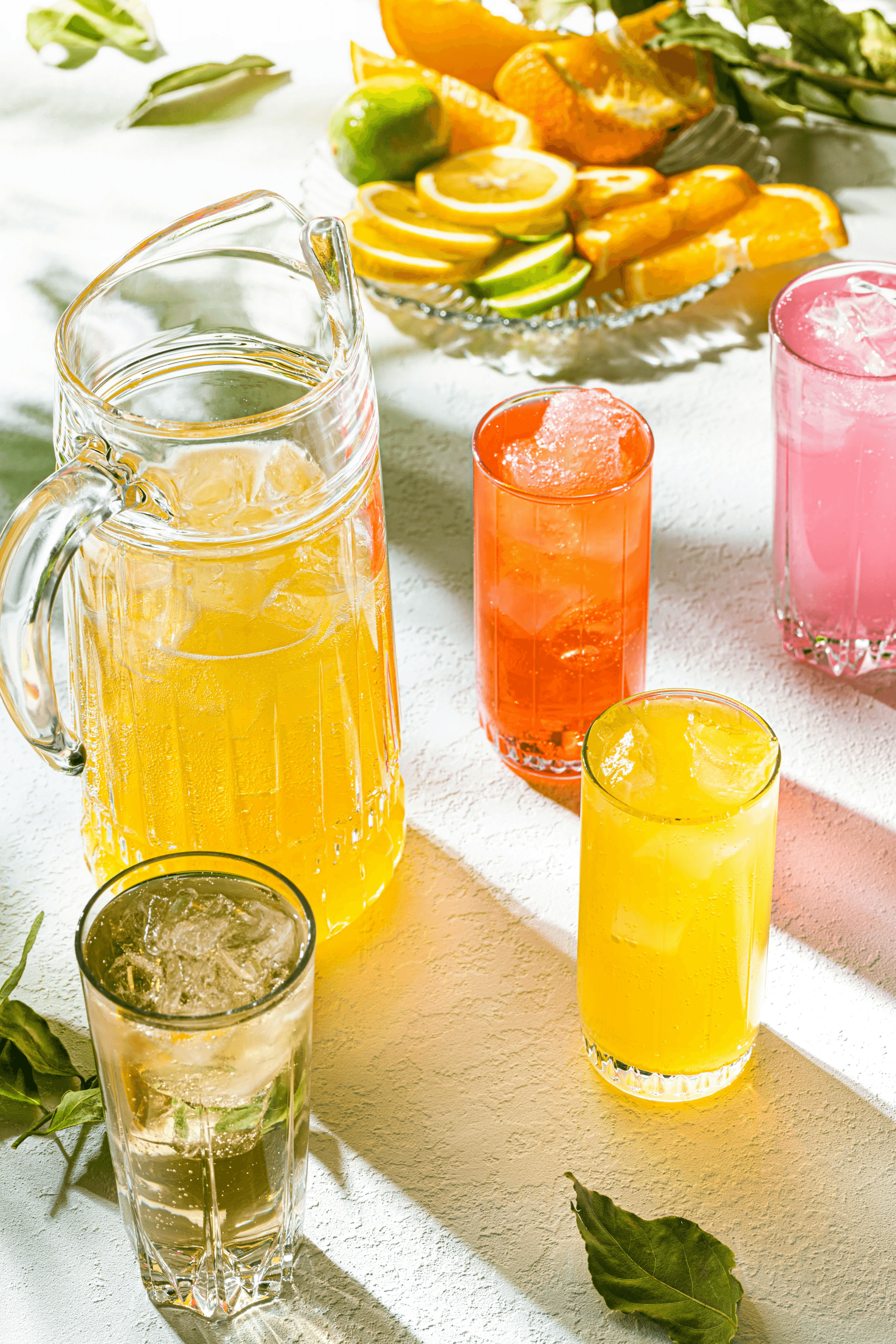 Brightly lit scene with a pitcher and glasses filled with colorful citrus drinks, surrounded by slices of lemon, lime, and orange on a textured white surface.