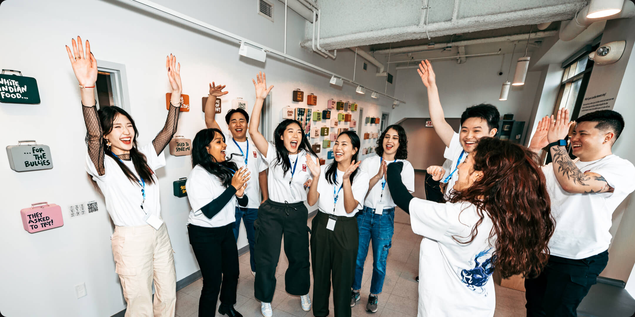 This image shows the TCP staff enthusiastically celebrating during their Lunar New Year Bash event. The group, dressed in white TCP shirts, is gathered in a hallway with various colorful signs on the wall behind them. They are laughing, cheering, and raising their hands in joy, capturing the festive and spirited atmosphere of the event.