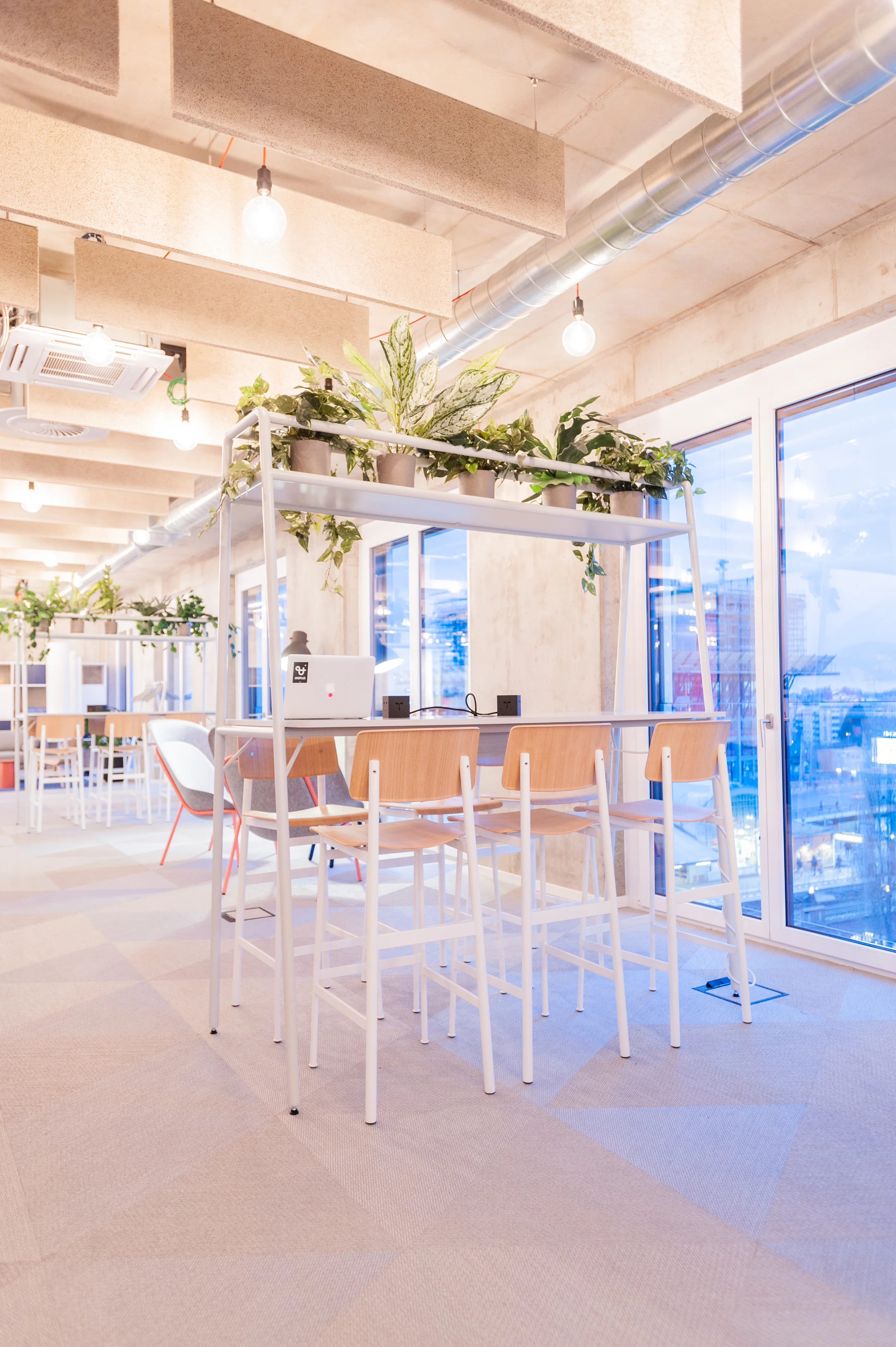 High table at OMIO with white frame, plywood bar stools and a plant shelf above, standing by floor-to-ceiling windows with an evening city view.