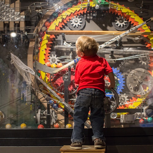 Un niño está de pie sobre un banco, observando de cerca una máquina colorida e intrincada con engranajes, pistas y bolas.