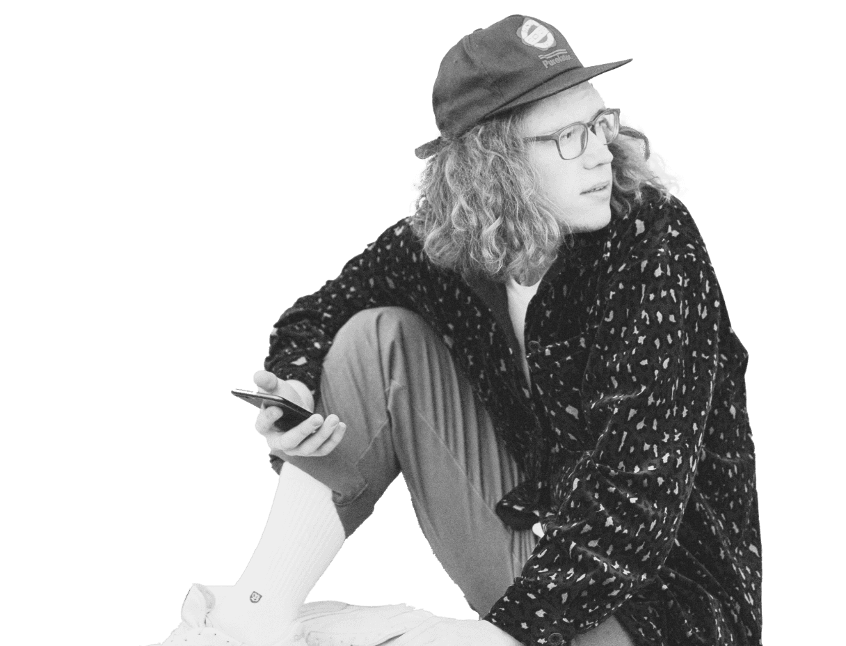 A person with long curly hair, wearing glasses, a patterned jacket, pants, a baseball cap, and sneakers, is sitting casually while holding a smartphone, conveying a relaxed and thoughtful demeanor, potentially indicative of using a trading demo account.