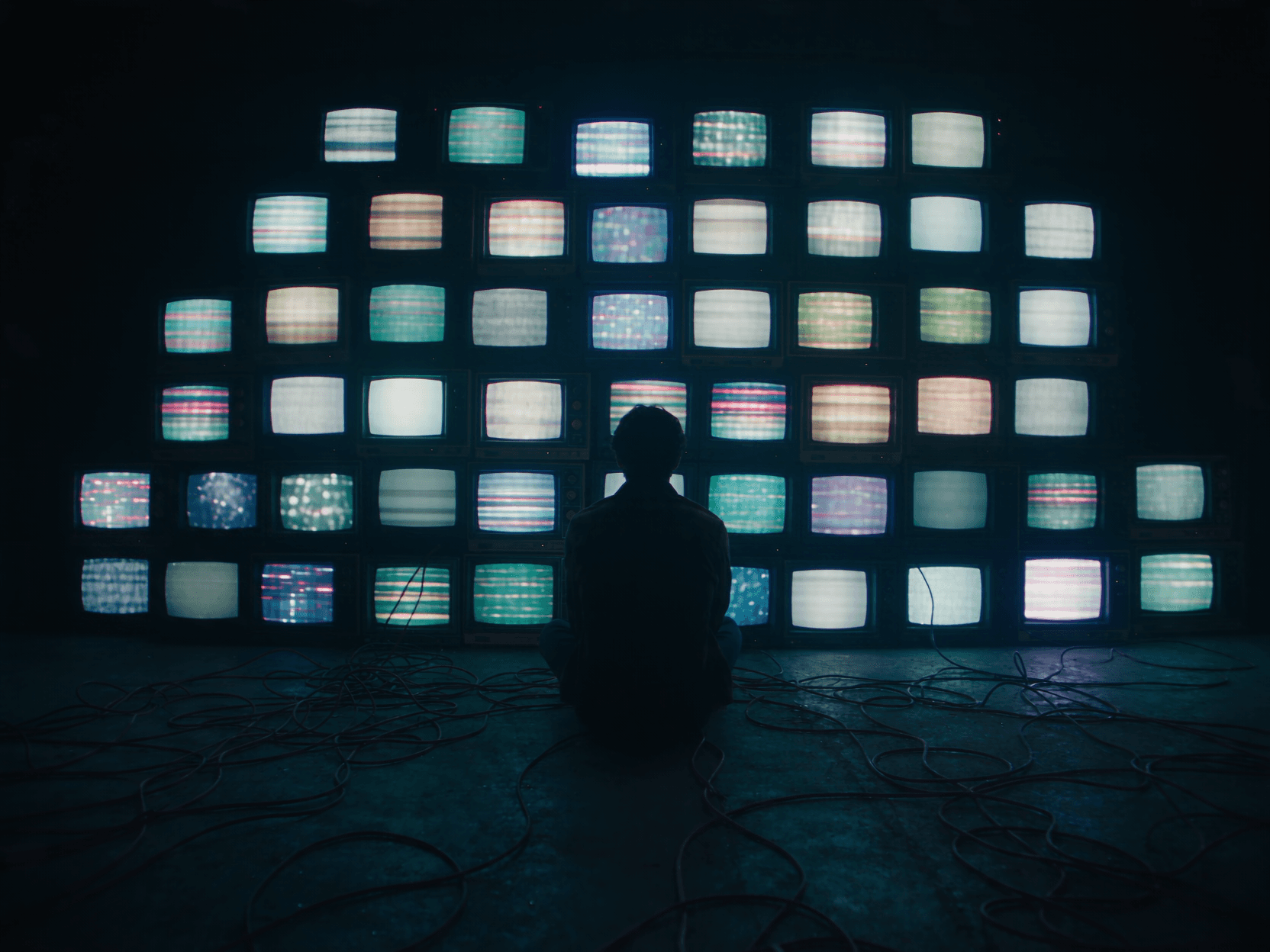 A man sits in the dark before a curved wall of old televisions showing static