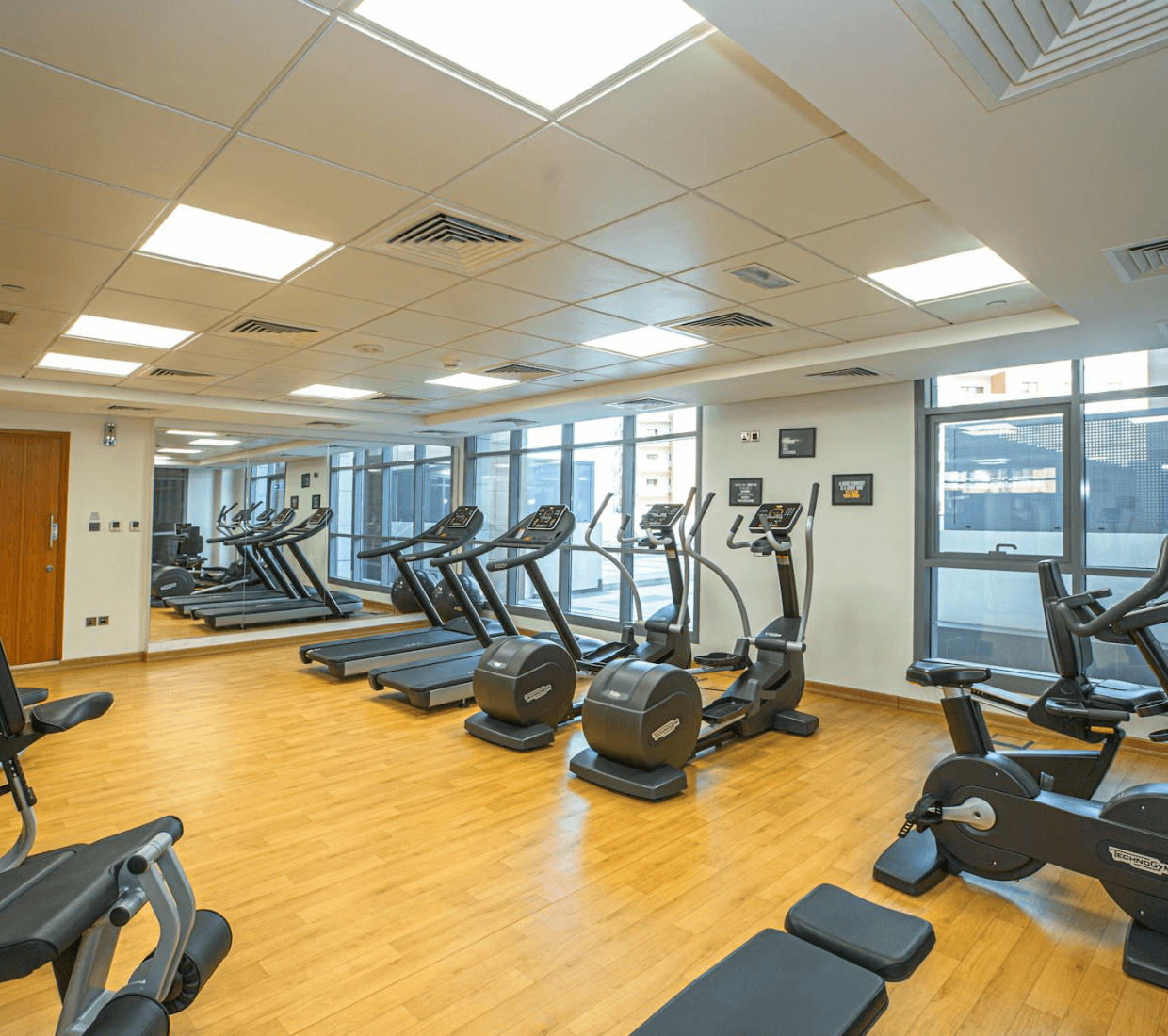 Bright gym with wood floors, treadmills, ellipticals, stationary bikes, and weight machines near windows and mirrors.
