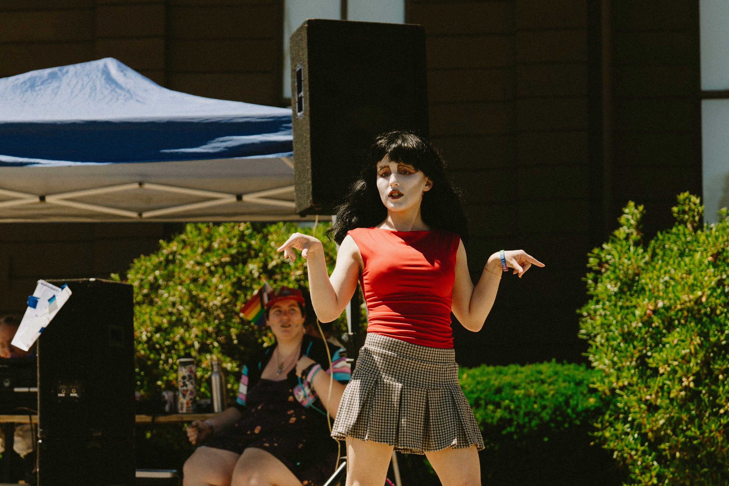 A drag performer in a black wig, red top, and plaid skirt performs mid-set at Middleboro PrideFest, speaker stacks rising behind.