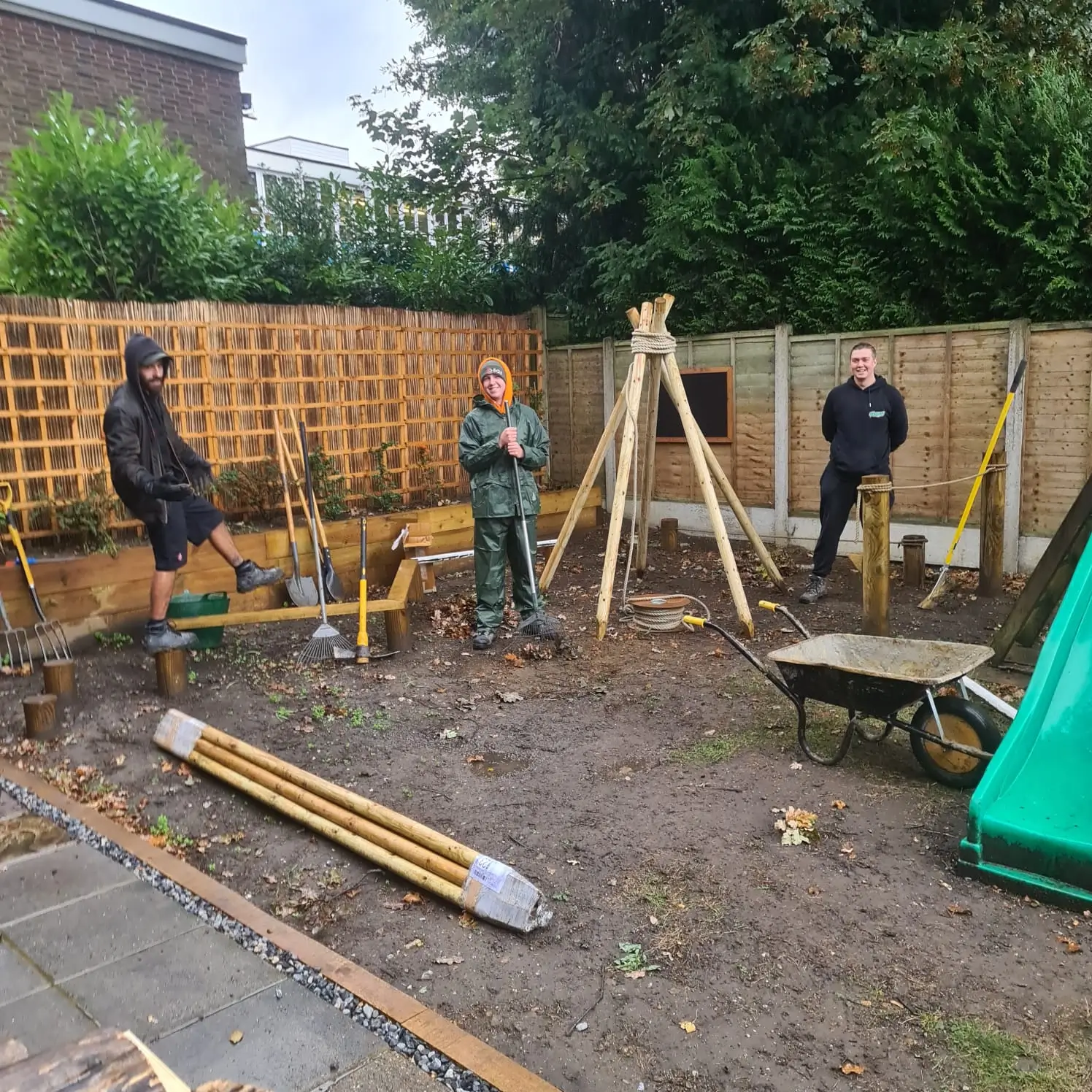 A group of people working in a yard with tools and materials, surrounded by wooden fencing and green vegetation.