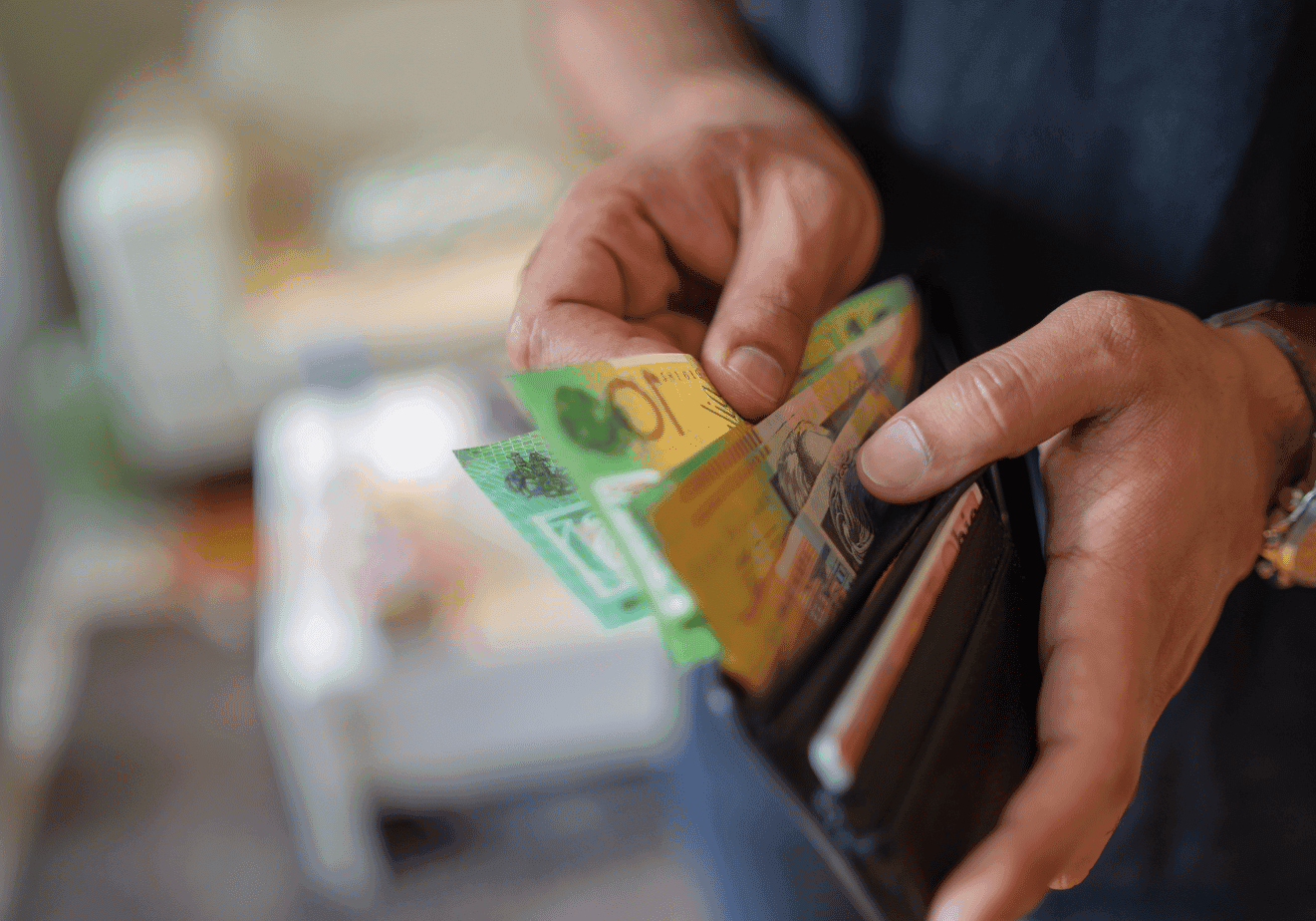 A person is holding a wallet open, counting Australian banknotes in various denominations, with a blurred living room background.