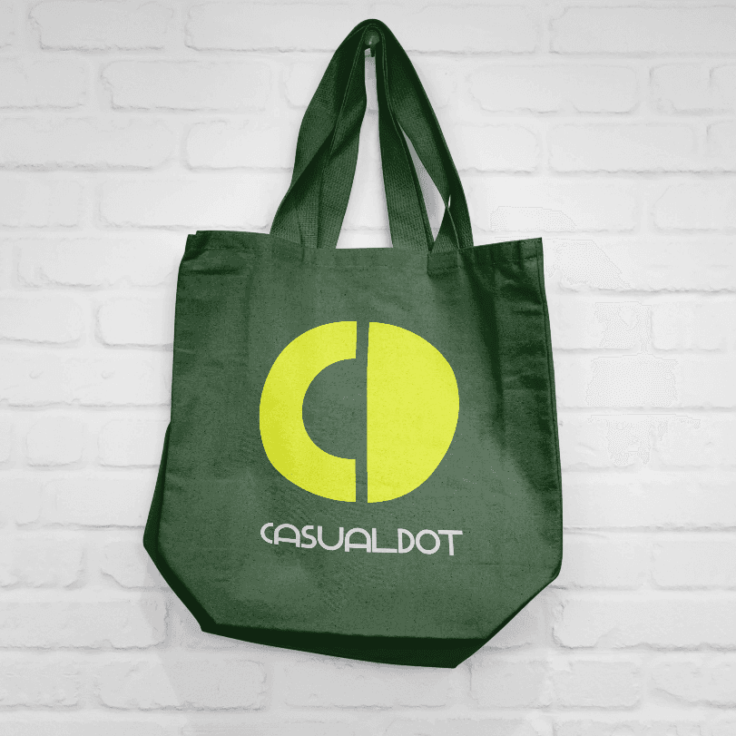 Green tote bag with yellow CD logo for Casual Dot, hanging on a white brick wall.
