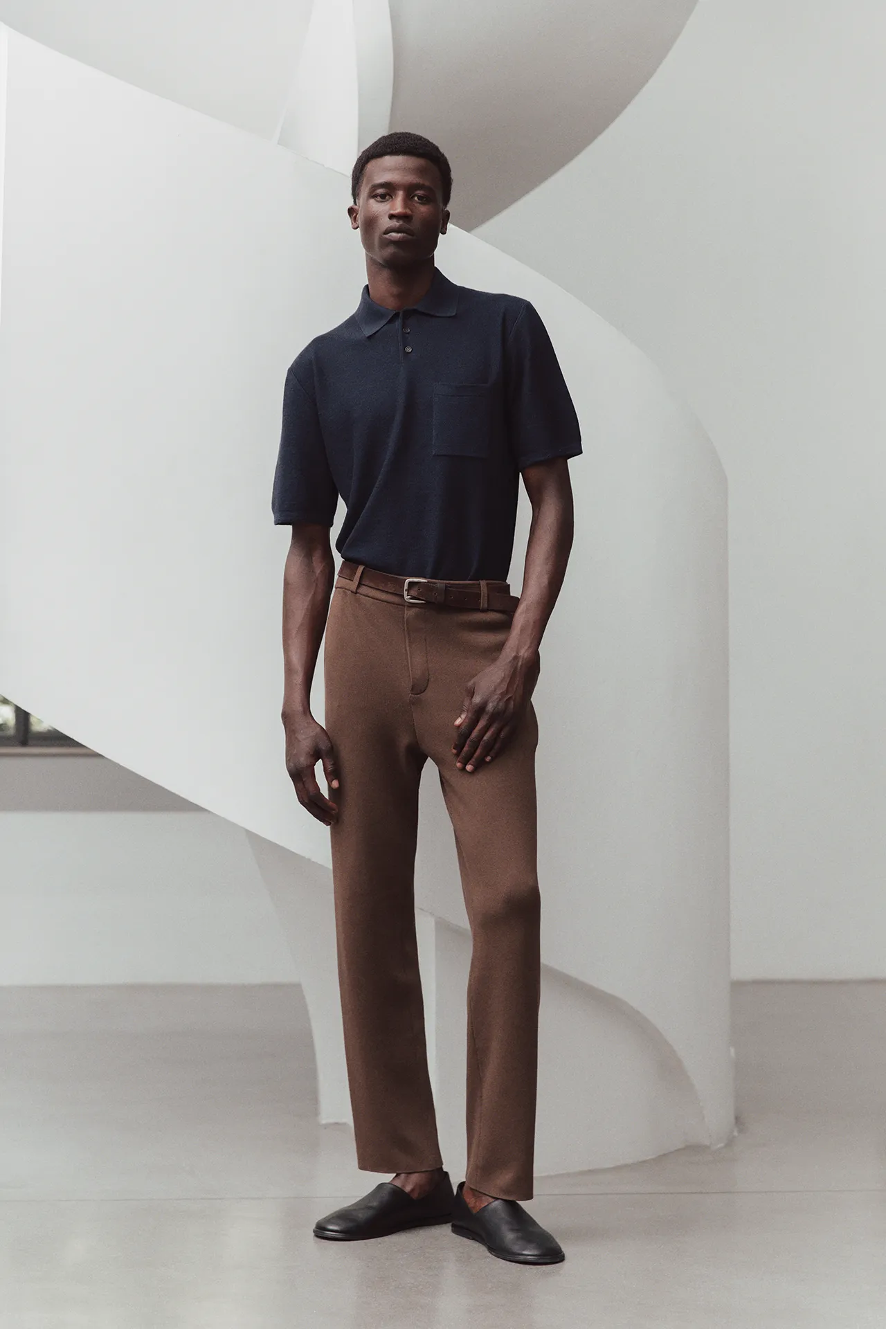 Full-length image of a model wearing the Aida Barni SS26 collection men's navy blue knit polo shirt paired with brown trousers.