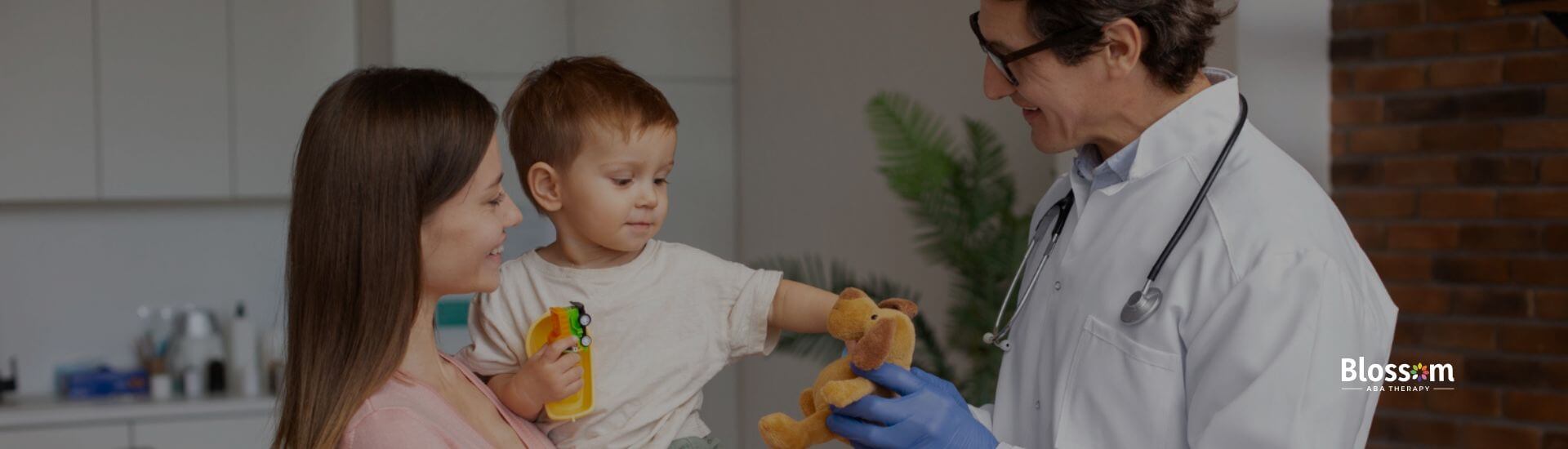 A doctor smiling and showing a teddy bear to a child with autism being held by their mother.