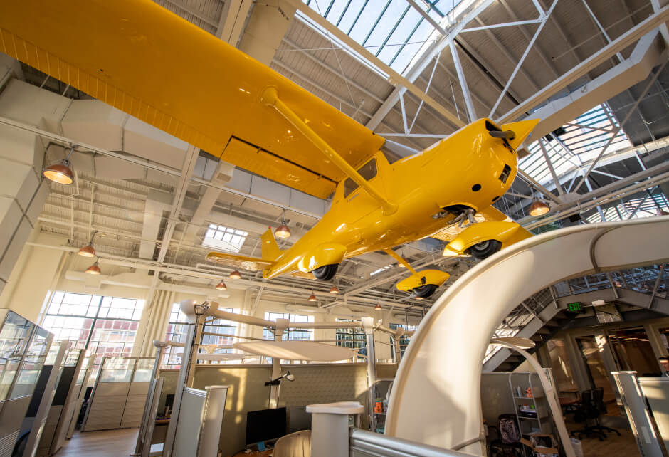 A bright yellow airplane is suspended from the ceiling inside a modern office space with open workstations and large windows.