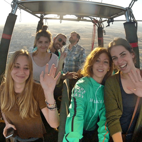 Five people smiling and waving inside a hot air balloon basket with a landscape visible in the background.
