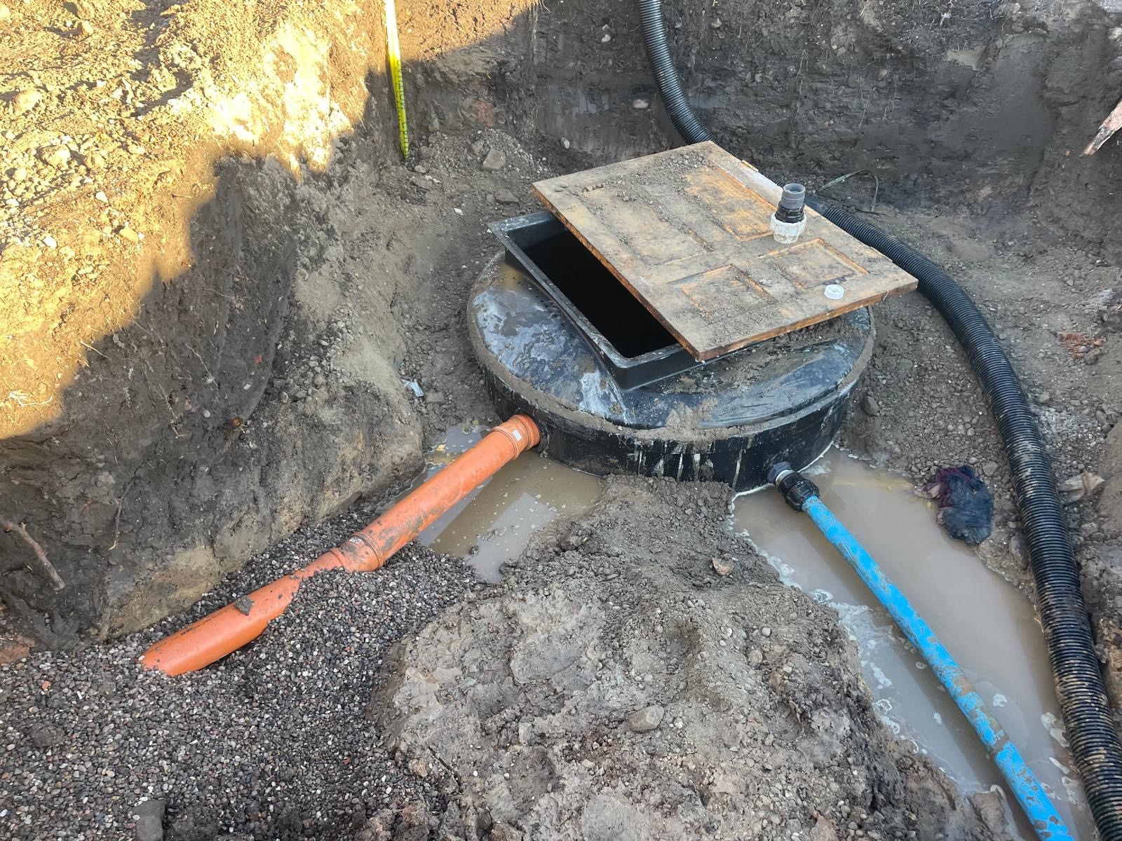 A circular manhole cover is partially uncovered in a dirt trench, with hoses and cables nearby.