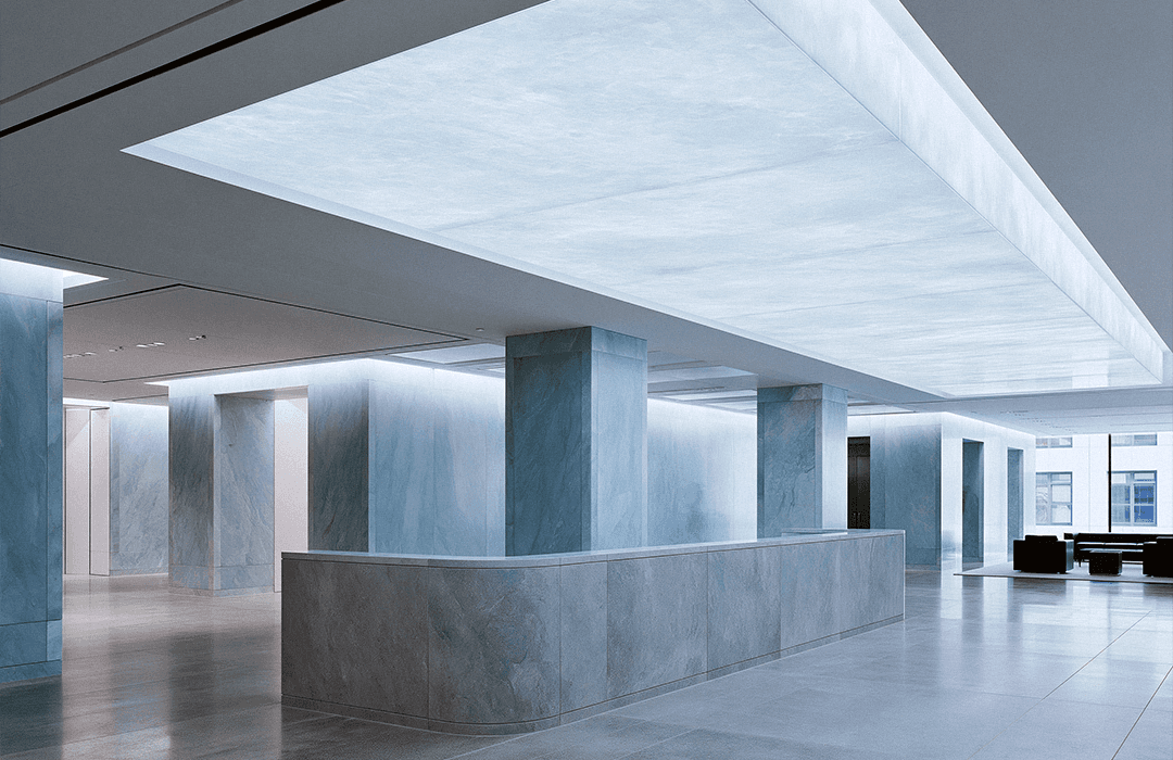 Fotografia do lobby amplo e minimalista de um edifício comercial moderno. O espaço possui colunas e balcão de recepção em mármore cinza e azul-claro, com teto iluminado por um grande painel translúcido, transmitindo sofisticação e tecnologia. Imagem de capa do blog da Inspira.