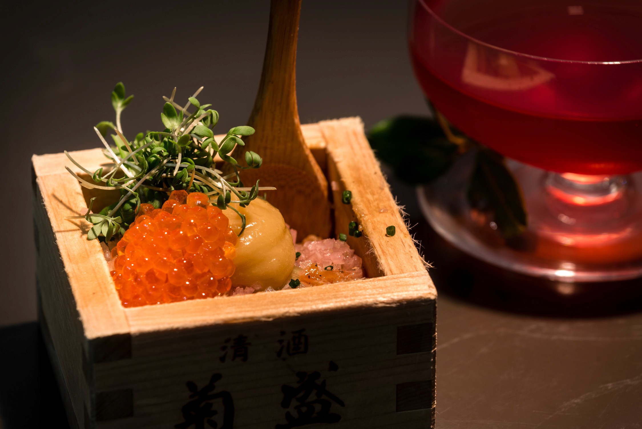 A wooden box filled with orange salmon roe, a yellow scallop, and microgreens sits next to a glass of red liquid.