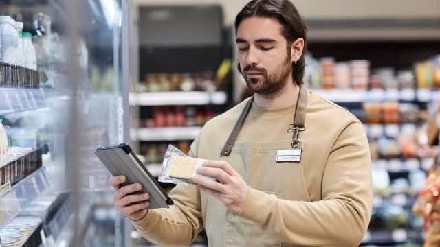 Homem de avental segurando tablet na mão direita enquanto analisa a embalagem do produto na mão esquerda, em ambiente de mercado.