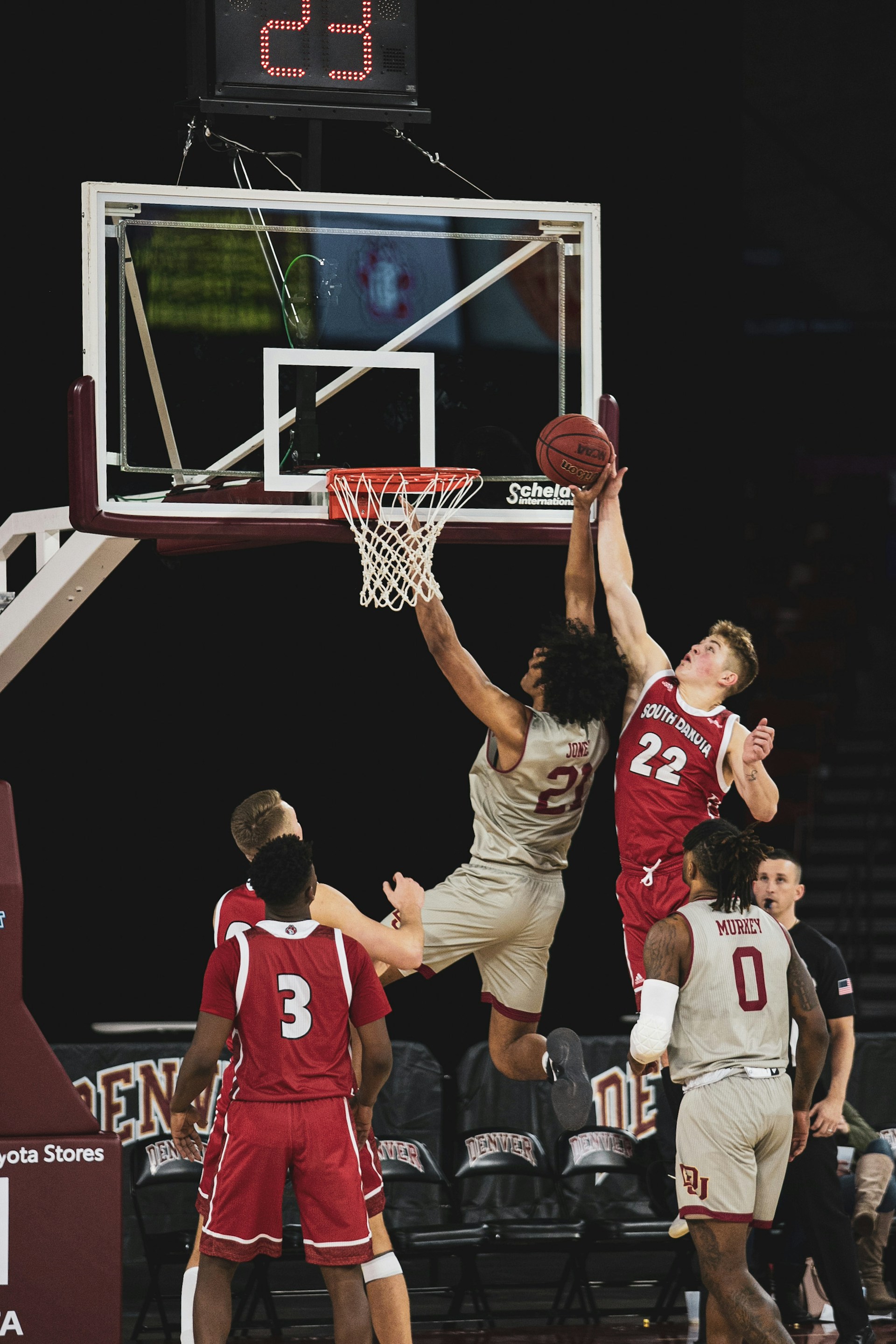 Two basketball players, one in grey and one in red #22, jumping high to contest a rebound near the hoop.
