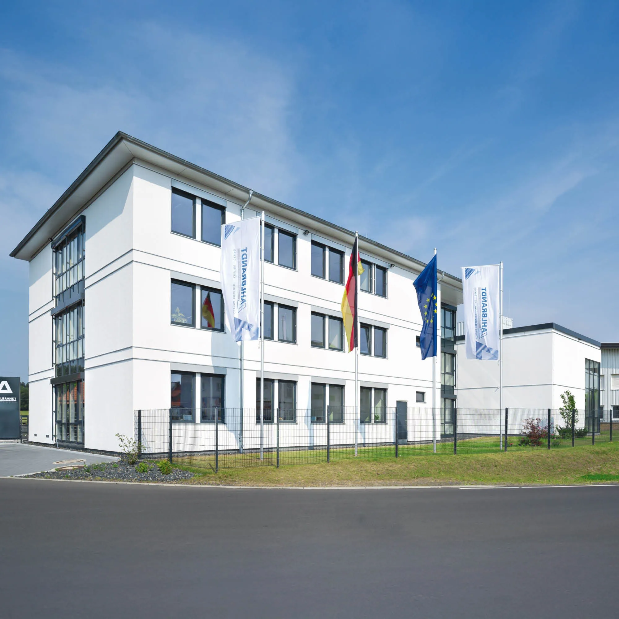 Mehrstöckiges weißes Verwaltungsgebäude mit vielen Fenstern, Flaggen von Deutschland und der EU im Vordergrund.