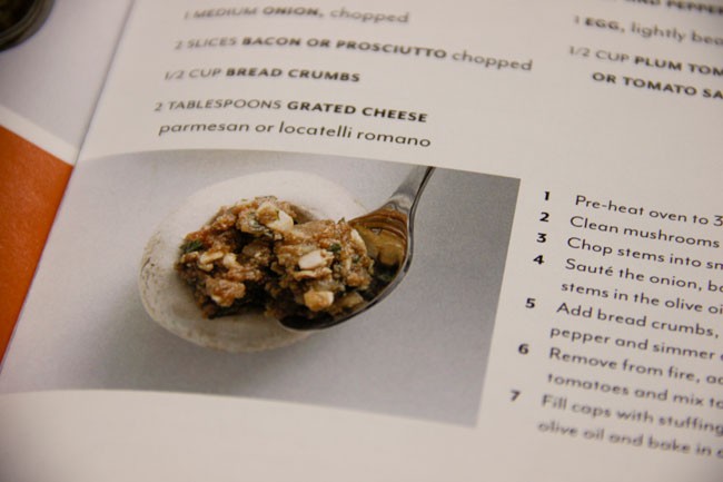 A close-up image shows a cookbook page with a recipe for pasta dish. The recipe includes ingredients like 1 medium onion, 2 slices bacon or prosciutto, 1/2 cup bread crumbs, 2 tablespoons grated cheese, parmesan or locatelli romano cheese, and instructions for preheating the oven to 350°F, cleaning mushrooms, chopping stems, adding bread crumbs, sautéing onion, and simmering in olive oil or tomatoes. The page is numbered 1 through 7. A photograph of a bowl filled with the prepared dish is centered on the page.