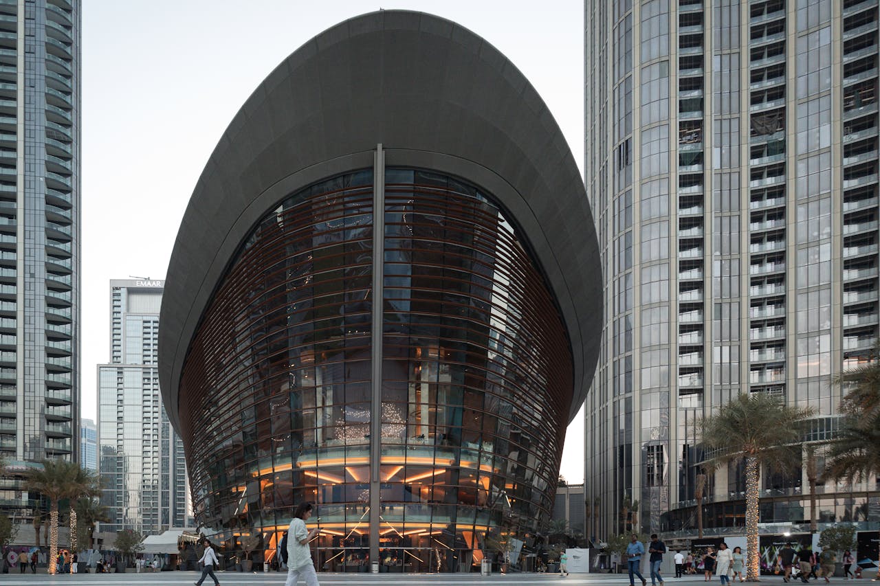 An oval-shaped glass building with dark curved exterior panels surrounded by tall towers.&nbsp;