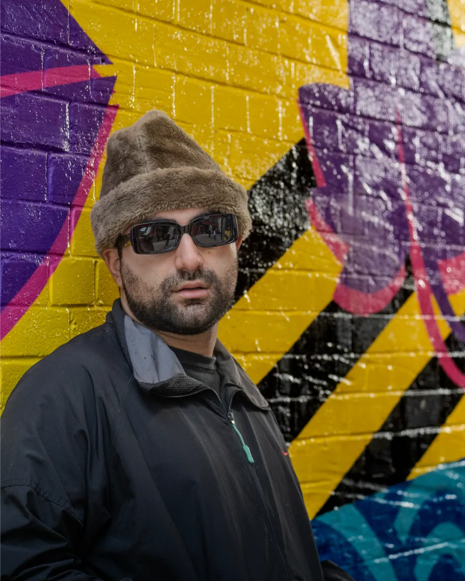 Aria posing against a vibrant yellow and purple mural off Coney Street, wearing a black windbreaker and dark sunglasses.
